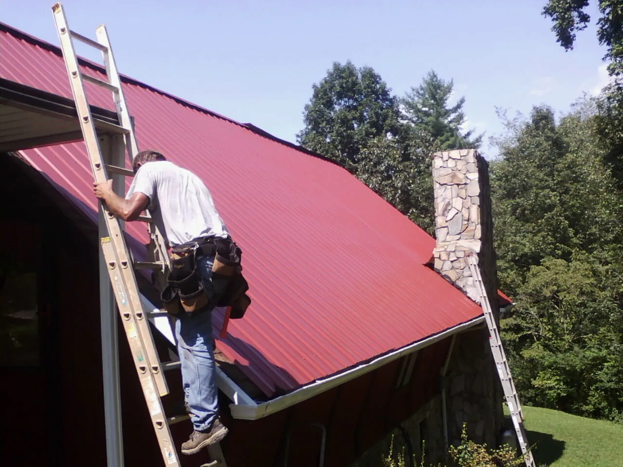 Roofer on a ladder beside a red metal roof and stone chimney — Soffit & Fascia Repair by Roofing Brothers in Allenspark